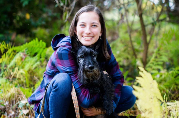 Smiling woman in a plaid shirt crouching in a forest, hugging a black dog.